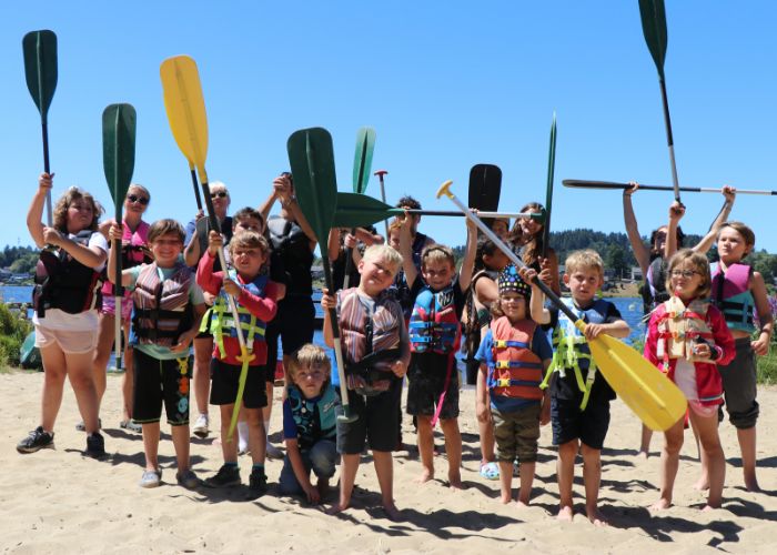 A group of campers standing on the shore holding canoe paddles up in the air